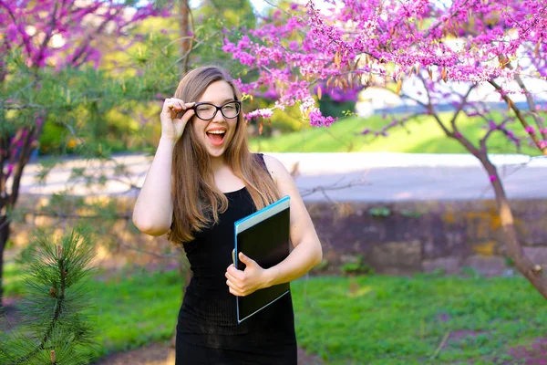 Studente intelligente femminile che tocca gli occhiali in giardino e in piedi con documenti in cartella nera . — Foto Stock