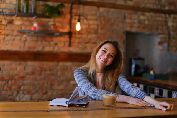 Fröhliches Mädchen am Tisch mit einer Tasse Kaffee und einem Notizbuch in der Mittagspause. — Stockfoto