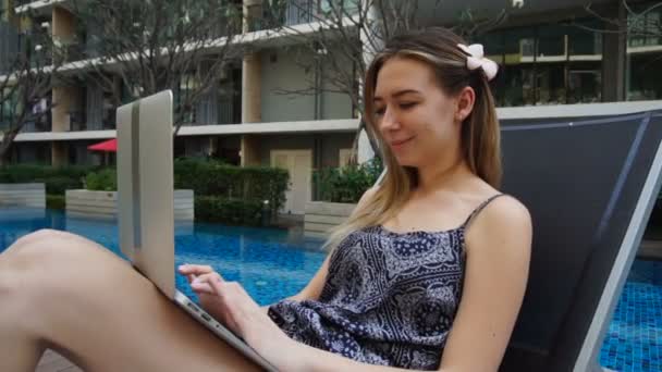 Femeie tânără întinsă pe un scaun de plajă și folosind laptopul lângă piscină — Videoclip de stoc