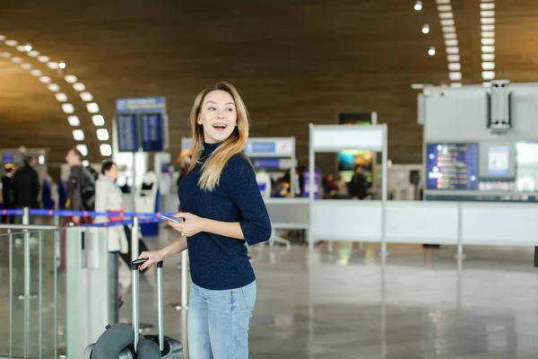 Girl walking in airport hall with smartphone and valise with neck pillow.