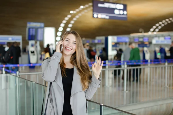 Kobieta rozmawia przez smartfon w hali lotniska, noszenie płaszcz szary i czarny worek, pokazano ok. — Zdjęcie stockowe