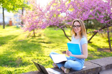Dizüstü bilgisayar ve çiçek açan park klasöründe gazetelerde oturan gülümseyen kız öğrenci.