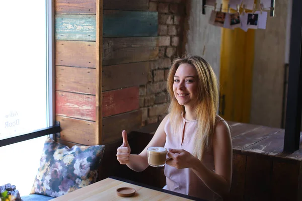 Młoda kobieta siedzi w pobliżu okna w przytulnej kawiarni i przytrzymując filiżankę cappuccino. — Zdjęcie stockowe