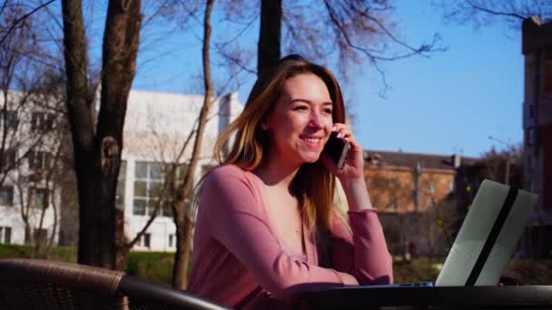 Молода жінка розмовляє по смартфону і переглядає ноутбук на свіжому повітрі з фоном блакитного неба . — стокове відео