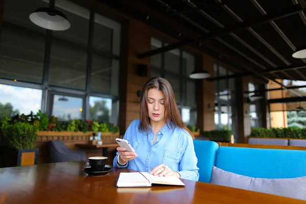 Девушка держит смартфон, читает блокнот и отдыхает в кафе с кофе . — стоковое фото