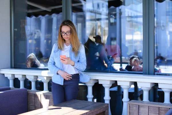 Chica bonita usando fuentes de Internet por smartfone y de pie en la cafetería . — Foto de Stock