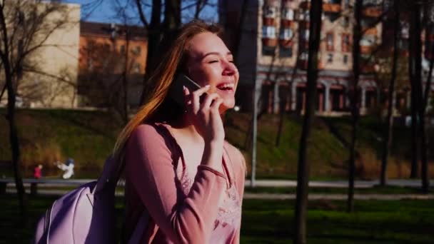 Fröhliche Studentin geht im Park spazieren und spricht in Zeitlupe mit dem Smartphone. — Stockvideo