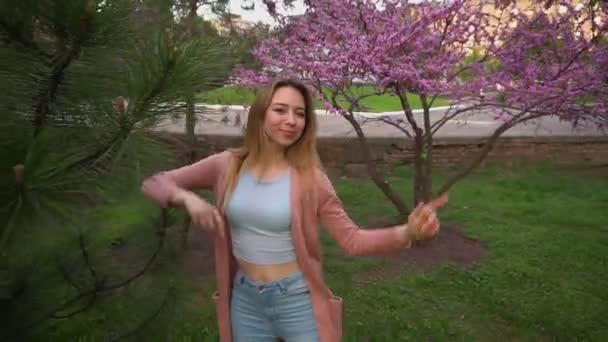 Jocund menina dançando perto de árvores florescendo no parque e soprando beijo . — Vídeo de Stock