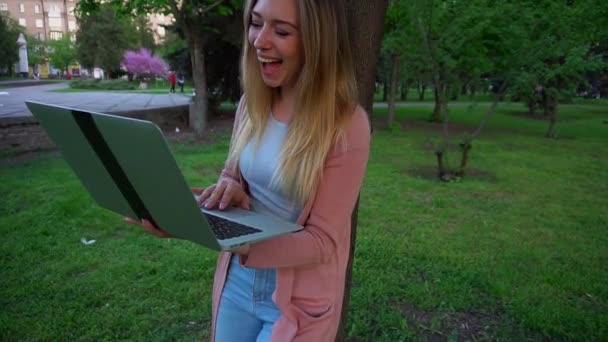 Jocund girl browsing by laptop and leaning on tree in spring park. — Stock Video