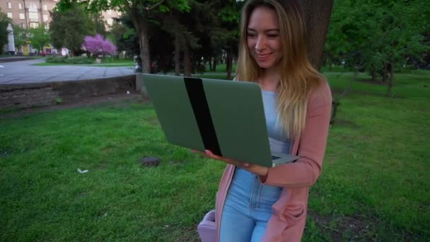 Mädchen plaudert mit Laptop im Park und lehnt mit zufriedenem Gesicht in Zeitlupe an Baum. — Stockvideo