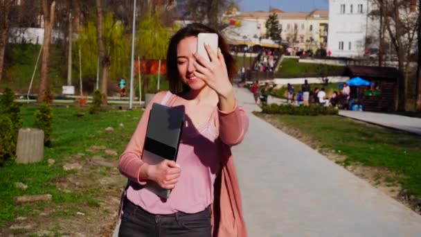 Красива дівчина робить селфі за допомогою смартфона з ноутбуком біля парку в повільному русі . — стокове відео