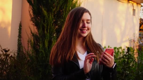 Mooi meisje bericht aan het typen door smartphone in roze geval in de buurt van groene planten. — Stockvideo