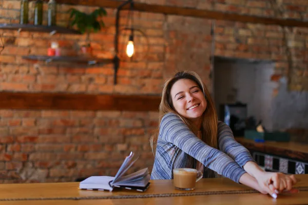 Zadowolony dziewczyna siedzi w pobliżu tabeli filiżankę cappuccino i notebook w przytulnej kawiarni z niezwykłym wnętrzu. — Zdjęcie stockowe