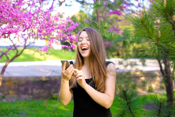 Meisje met nieuwe smartphone in park met bloeiende bomen achtergrond lachen. — Stockfoto