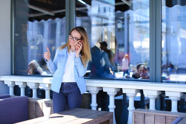 Estudiante de pie en el café de la calle, mostrando los pulgares hacia arriba y hablando por teléfono inteligente . — Foto de Stock