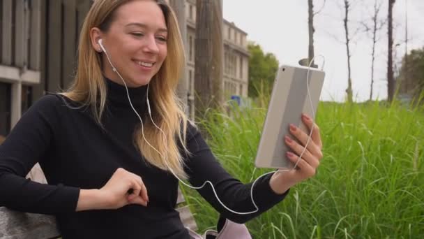 Mujer hablando por tableta y en auriculares con videollamadas, sentada en el banco y mostrando los pulgares hacia arriba en cámara lenta . — Vídeos de Stock