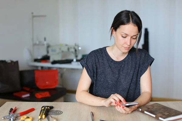Hermosa artesana sentada en el taller y usando un teléfono inteligente, artículos de cuero hechos a mano . — Foto de Stock