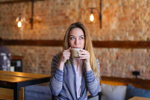 Zadowolony dziewczyna siedzi w pobliżu tabeli filiżankę cappuccino w przytulnej kawiarni z niezwykłym wnętrzu. — Zdjęcie stockowe