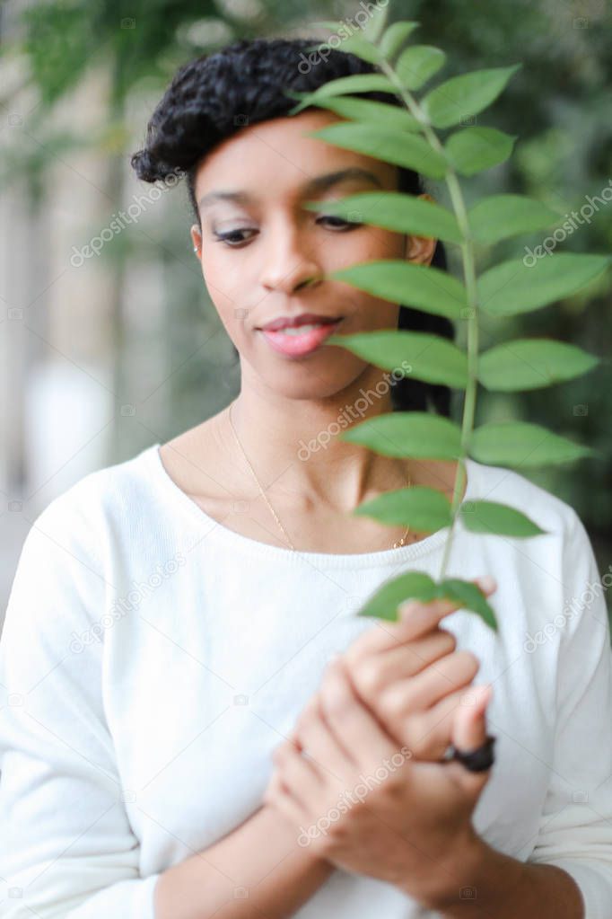 Black half american nigerian girl standing with green leaf, having bangs and wearing white blouse.