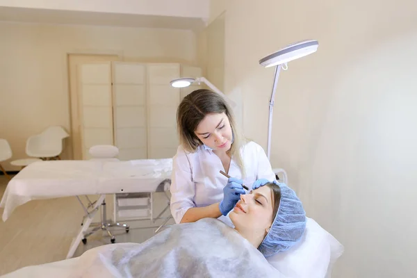 Kosmetikerin macht Make-up mit Augenbrauen-Tätowierstift für Frau. — Stockfoto