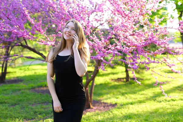 Nettes Mädchen, das im Frühlingspark spaziert und mit dem Smartphone spricht. — Stockfoto