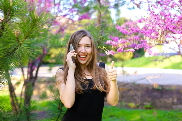 Mujer hablando por teléfono inteligente y mostrando los pulgares hacia arriba en el parque de primavera . — Foto de Stock
