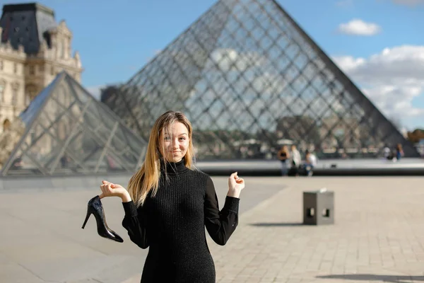 Boso dziewczynę stojącą w pobliżu Luwru i szkło pyramind w czarnej sukience w Paryżu z buta w ręku. — Zdjęcie stockowe