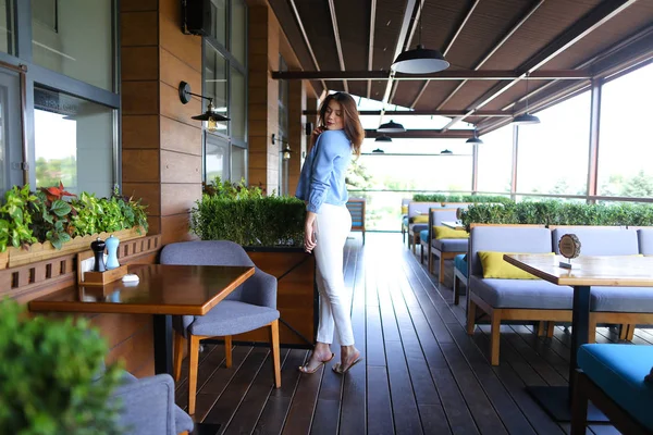 Menina de pé no café e mantendo smartphone, vestindo camisa jeans e calças brancas . — Fotografia de Stock