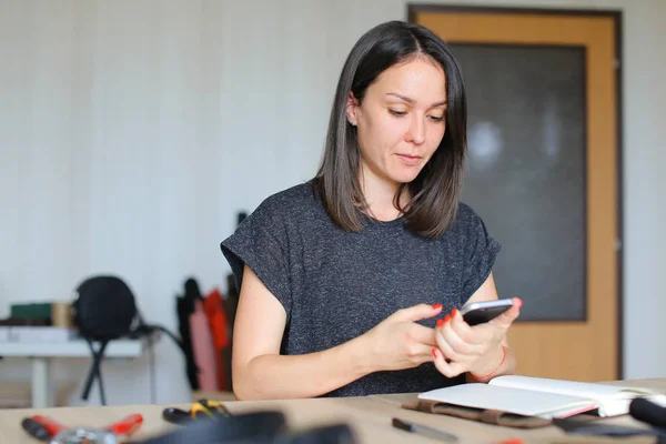 Chica creando portátil de cuero hecho a mano y utilizando el teléfono inteligente para nuevas ideas . — Foto de Stock