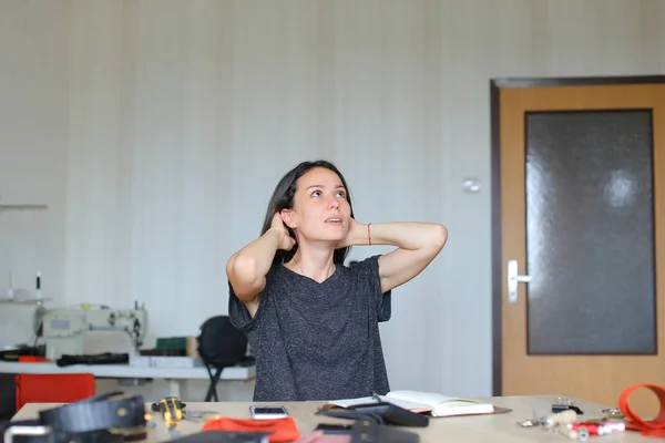 Mujer joven sentada en el taller de cuero y haciendo portátil hecho a mano, teléfono inteligente en la mesa . —  Fotos de Stock