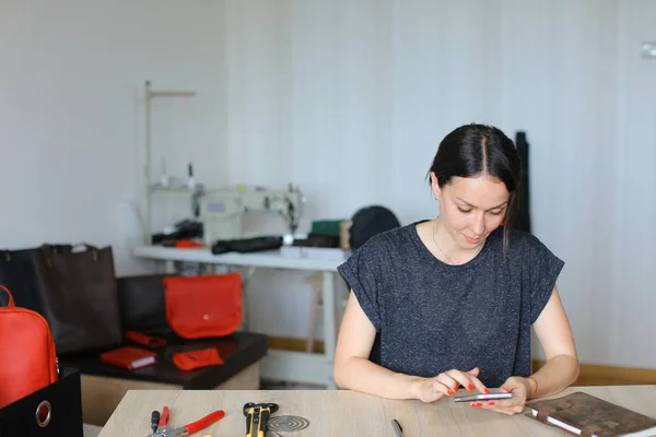 Artesana sentada en el taller y usando smartphone, artículos de cuero hechos a mano . — Foto de Stock