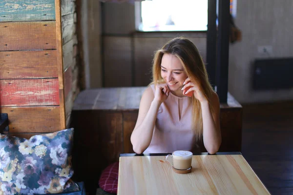 カプチーノのガラスのカップと木製のテーブルの近くに居心地の良いコーヒー家で座っていると、スマート フォンを使用して若い女性. — ストック写真