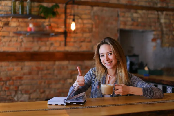 Uśmiechnięta dziewczyna siedzi w pobliżu tabeli z notebooka i przytrzymując filiżankę cappuccino. — Zdjęcie stockowe