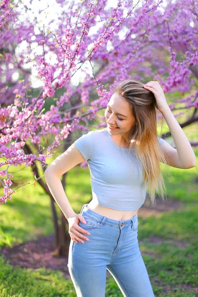 Lächelnde blonde Frau steht im Park mit Blütenhintergrund. — Stockfoto