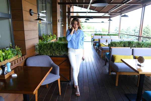 Mulher elegante em pé no café com smartphone e vestindo camisa jeans e calças brancas . — Fotografia de Stock