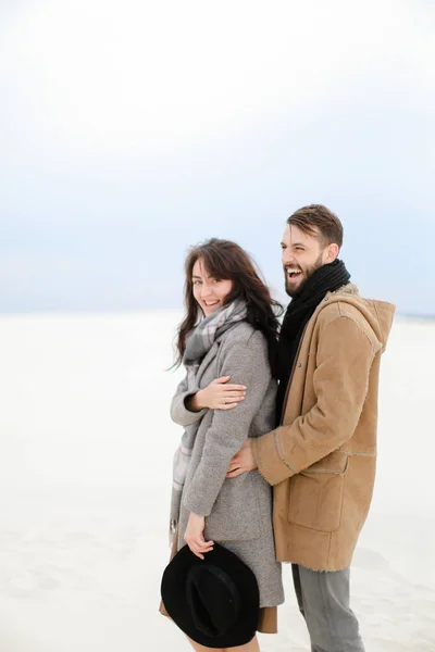 Homem caucasiano feliz abraçando a pessoa feminina vestindo casaco cinza e cachecol, fundo monofônico de inverno branco . — Fotografia de Stock