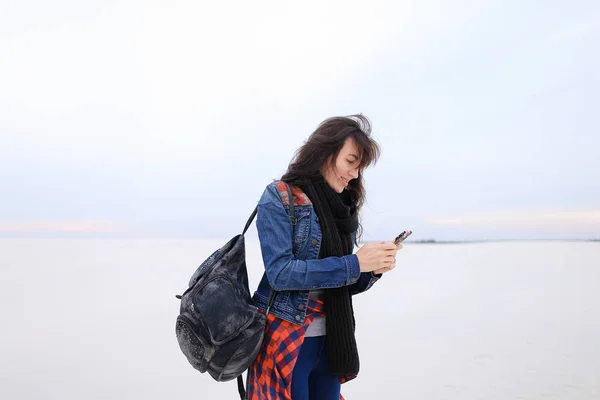 Elegante chica con pantalones vaqueros y bolso, usando teléfono inteligente y de pie en el fondo blanco de invierno . —  Fotos de Stock
