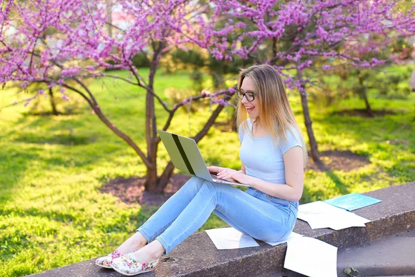 Étudiante joyeuse se préparant à l'examen avec ordinateur portable et papiers dans le parc en fleurs . — Photo