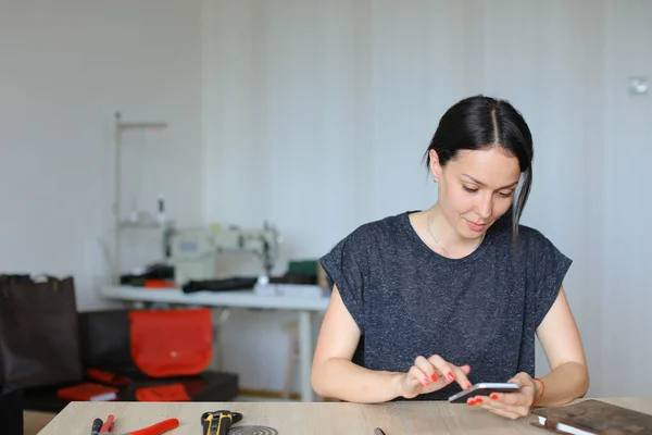 Καυκάσιος craftswoman κάθεται στο ατελιέ και χρησιμοποιώντας το smartphone, χειροποίητα δερμάτινα είδη. — Φωτογραφία Αρχείου