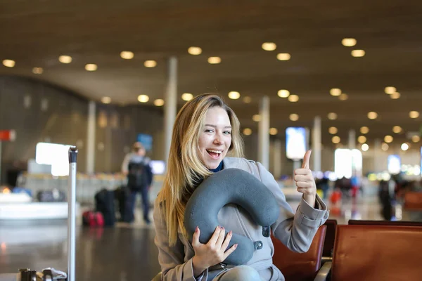 Nice woman sitting with neck pillow and valise in airport waiting room, showing thumbs up. — Stock Photo, Image