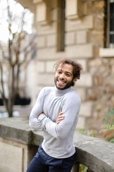 Preto sorrindo menino vestindo cinza gola alta camisola de pé perto de concreto corrimão perto de tijolo parede fundo . — Fotografia de Stock