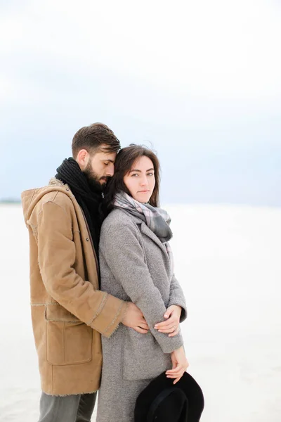 Junger Mann umarmt hübsche Frau in grauem Mantel und Schal vor weißem Winterhintergrund. — Stockfoto