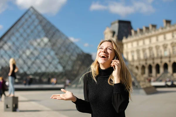 Pretty girl standing near Louvre and glass pyramind in black dress in Paris, talking by smartphone. — Stock Photo, Image