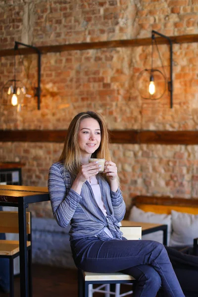 Zadowolony dziewczyna siedzi w pobliżu tabeli filiżankę cappuccino w przytulnej kawiarni z niezwykłym wnętrzu. — Zdjęcie stockowe