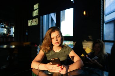 Smartphone kullanarak ve güneş ışınları kafede oturan genç kadın.