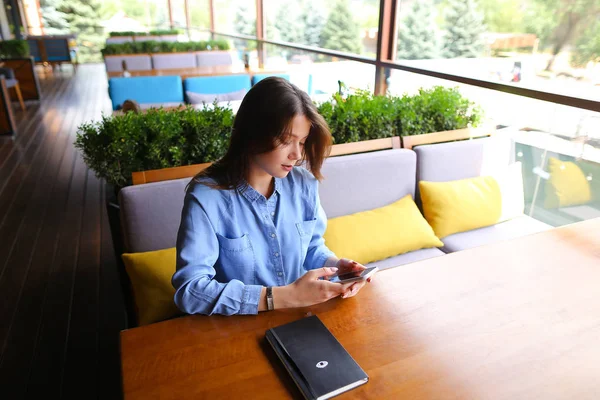 Hermosa estudiante sentada en la cafetería con portátil en la mesa y el uso de teléfono inteligente . — Foto de Stock