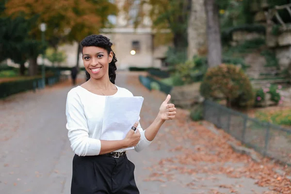 Afro americký mladá podnikatelka chůzi s doklady v podzimním parku, ukazuje palec. — Stock fotografie