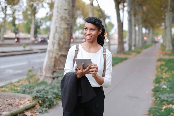 Black happy female student walking with tablet near street with trees.