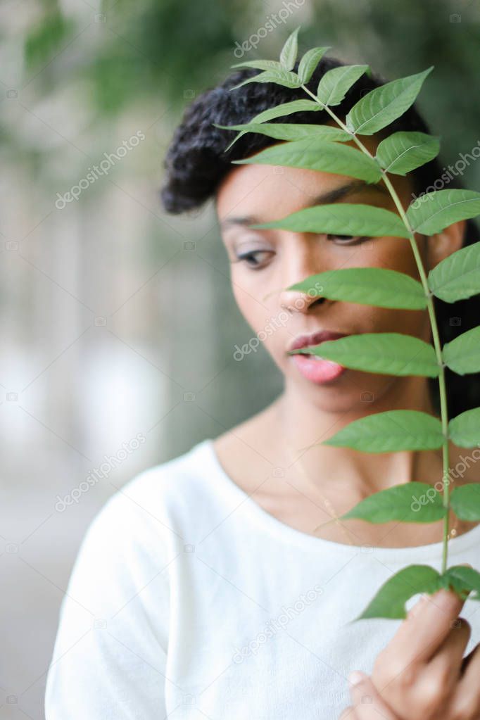 Close up face of black girl keeping green leaf, having bangs and wearing white blouse.