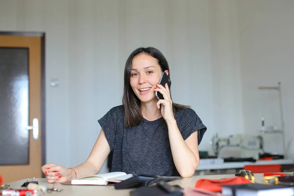 Alegre persona femenina sentada en el taller de cuero y haciendo cuaderno y billeteras hechas a mano, hablando por teléfono inteligente — Foto de Stock